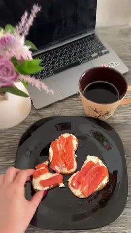 A close-up video shows a black plate with three open-faced sandwiches on it. Each sandwich consists of a slice of bread topped with cream cheese and slices of smoked salmon. A hand with dark pink nail polish reaches for one of the sandwiches. To the right of the plate is a tan mug filled with a dark liquid, likely coffee, with a floral design. Behind the mug and plate is a laptop computer. To the left of the laptop is a vase with a pink and purple floral arrangement. The background appears to be a wooden surface.