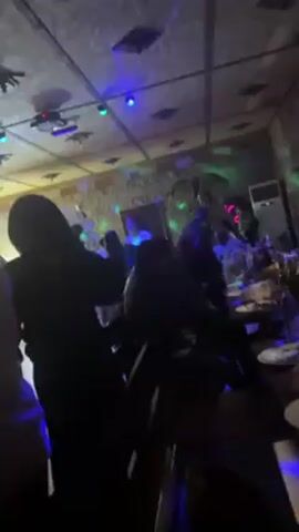 The video shows a crowded dance floor in a dimly lit venue. A DJ is visible on a stage at the far end of the room, and the ceiling has several disco lights. People are dancing and interacting with each other, but their faces are not easily discernible due to the lighting conditions. There's an air conditioning unit on the right side of the room.