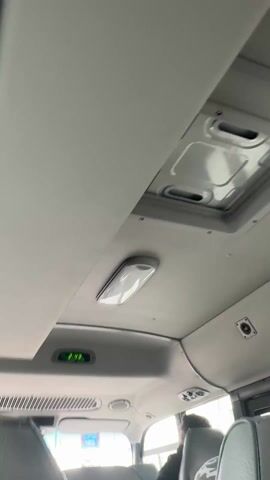 The camera points toward the ceiling inside a bus, showing an electronic clock that reads 2:43, as the bus appears to be in motion. There are lights and vents in the ceiling. The tops of passengers’ heads and the backs of seats are visible in the lower part of the frame. The seats are light gray. The ceiling is off-white.