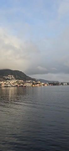The video shows a harbor town along a body of water, possibly a fjord or bay. The town is nestled against a hill that slopes upward, partially obscured by low-hanging clouds. The town's buildings appear to be a mix of residential houses and small commercial structures.  A number of boats are docked in the water. The overall light suggests early morning or late afternoon with diffuse sunlight.
