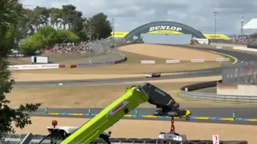Here is a concise description of the video:

The video shows a race at a racetrack. In the background, a "Dunlop" sign is visible. A green CLAAS crane is in the foreground, lifting a white object. A race car with a white, grey, and lime green color scheme is seen speeding by. There are crowds of people along the track. A "Cadillac" banner is visible as a blue and green race car speeds past.