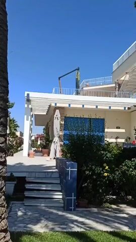 A video pans across the exterior of a white, multi-story building with blue accents. The building has multiple balconies with white railings and blue trim. There is a set of stairs leading up to a covered patio area with white columns. Lush green foliage and flowering bushes, some with red flowers, are visible around the building. A paved walkway leads through a grassy area. A white car is parked on a driveway in the background. The sky is clear and blue.