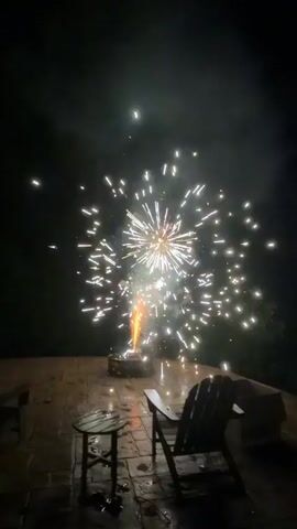 The video captures the momentary ignition and detonation of fireworks in a backyard setting at night. The fireworks are set off from a metal structure on a stone patio, which is wet, suggesting recent rain. In the foreground, two Adirondack chairs and a small round wooden table are visible. The fireworks emit a bright white light and sparks, illuminating the dark sky and casting a glow on the surrounding environment. The visual experience is brief, as the fireworks ignite and explode in rapid succession within a few seconds.