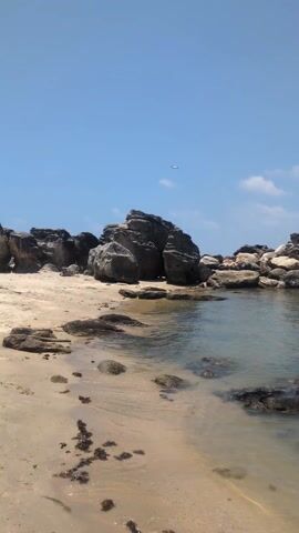 The video shows a beach with large, dark rocks scattered across the sand and in the water. The water is calm and clear. The sky is a bright blue with a few scattered white clouds. An airplane flies in the distance. The camera is stationary.