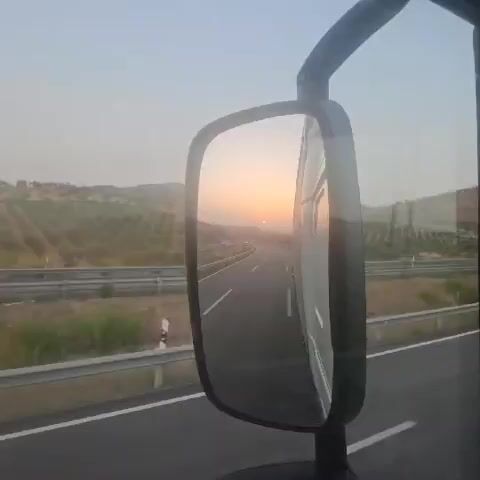A video taken from the side mirror of a vehicle shows a highway winding through a hilly landscape at sunset. The sun, a bright orange orb, is visible low on the horizon, casting a warm glow over the scene. The road ahead is empty except for a few cars, their headlights on. The landscape is dotted with trees and dry vegetation. The sky is a pale orange, gradually fading to a lighter hue towards the top. The video appears to be shot from a moving vehicle, as indicated by the slight blur and the reflection in the mirror. The overall mood is serene and picturesque.