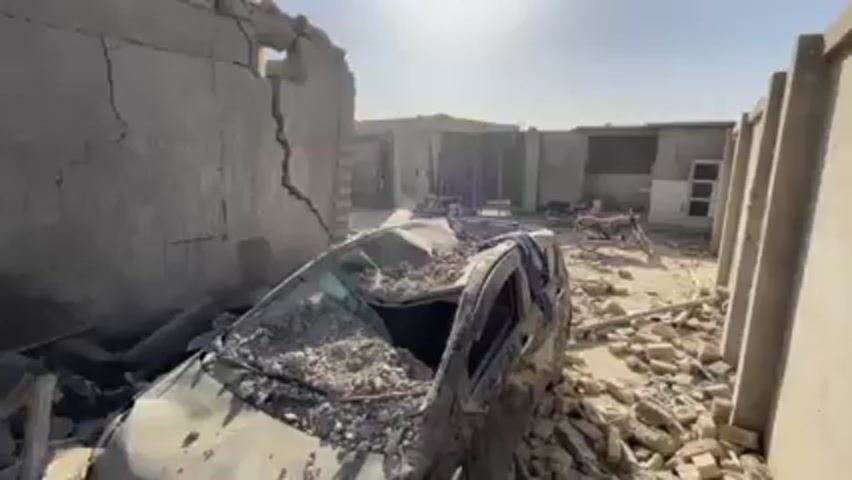The scene depicts a destroyed environment, possibly following an explosion or earthquake. A heavily damaged car is buried under rubble, with debris covering its hood and windshield. Adjacent to the vehicle, a building shows significant structural damage, including a large crack in its wall and missing sections. In the background, a group of people are gathered, seemingly observing the devastation. The setting appears to be a rural or semi-arid area, under a clear sky.