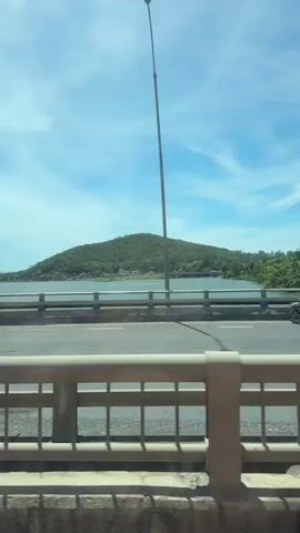 The video captures a view from a moving vehicle, looking across a bridge and towards a scenic landscape. In the foreground, the concrete structure of the bridge railing is visible, with its light-colored horizontal bars and vertical supports. The road surface of the bridge is also seen, with white lane markings.

Beyond the bridge, a body of water is present, with a distinctly shaped, green, tree-covered hill rising from it. The hill has a rounded peak and slopes down towards the water. The shoreline is lined with lush green trees, including palm trees. The sky is bright blue with scattered white clouds.

As the vehicle moves, motorcycles pass by on the bridge. In one instance, a person is seen riding a motorcycle on the bridge. Further down the road, another motorcycle is visible. There are also glimpses of traffic signs and streetlights along the roadside. The overall impression is a sunny day with a picturesque view of a coastal or lakeside environment.