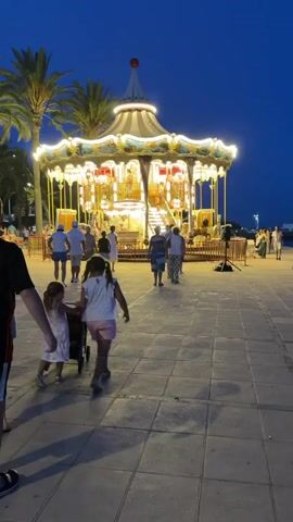 The video shows a carousel at dusk. A group of people are walking on a paved area in front of it. The carousel is lit with many lights and has several levels. Two children in the foreground are walking towards the carousel, one pulling a small cart. The sky is dark blue, indicating it is night or late evening. Palm trees are visible to the left of the carousel.