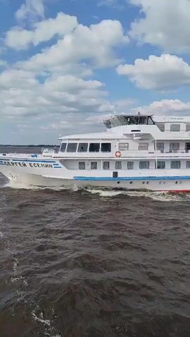 A white passenger ship is moving through dark, choppy water under a blue sky with white clouds. The ship has multiple decks with many windows and a white superstructure with a bridge at the front. The ship has a red and blue stripe along its hull. The video is filmed from a slightly elevated perspective, possibly from another boat.
