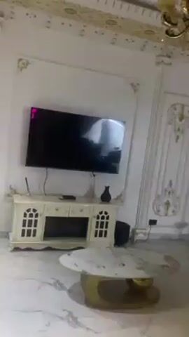 The video shows a lavishly decorated living room with white and gold accents. A flat screen television is mounted on the wall above a white cabinet. The room features a marble floor, an ornate coffee table with a gold base, and decorative wall paneling with gold detailing. A chandelier hangs from the ceiling. Various pieces of furniture are seen, including a large black chair, a small vanity stool, and a gold-colored sofa. There are no people visible in the video. The camera pans slowly around the room, showcasing the interior design.