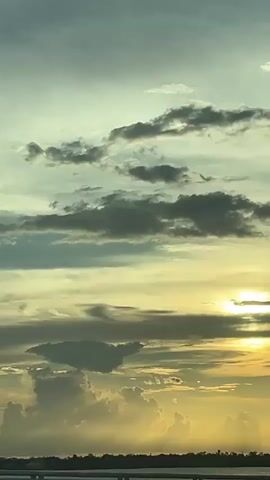 The video shows a sunset over a body of water, possibly a river or lake, with a bridge in the foreground. The sky is filled with dramatic clouds, some dark and stormy, others illuminated by the setting sun, creating a mix of blues, grays, and yellows. The sun itself is partially obscured by clouds, casting a warm, golden glow. The landscape on the far side of the water is silhouetted against the sky, with a few scattered trees visible. The overall mood is serene and majestic, with a sense of vastness and natural beauty.