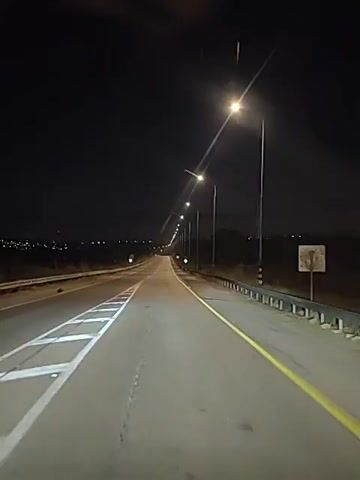 The video shows a nighttime view of a highway from the perspective of a vehicle in motion. Streetlights line the right side of the road, illuminating the asphalt. The road ahead curves gently upwards, disappearing into the darkness. The left side of the road has a white dashed line, and the right side has a solid yellow line. The sky is dark and clear, with a few distant lights visible on the horizon. The overall impression is one of a lonely, nocturnal journey.