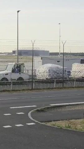 A white, enclosed utility vehicle drives along a road. Behind it, a line of cargo containers is visible. The containers are wrapped in white plastic and are stacked on flatbed trailers. A chain-link fence separates the road from the cargo. In the background, an airplane with a white fuselage and blue and white markings on its tail can be seen. The sky is overcast.