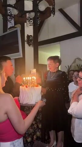 A woman in a black dress with pearls is surrounded by people and a birthday cake with lit candles. She is smiling and touching her ears as someone holds a phone to record the moment. The cake is white with red fruit on top and has seven lit candles. The environment appears to be indoors with wooden beams and lights visible.