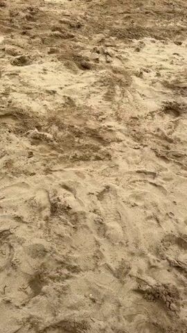 The video opens with a close-up of light brown sand, showing footprints and slight undulations. The lower legs and feet of a person in black shorts and a white top are visible at the top of the frame, standing on the sand. As the camera pans up, a man in a white polo shirt, black shorts, and a white baseball cap is seen standing on the sand with his hands on his hips, facing towards a long, open-sided structure with a thatched roof. Underneath the thatched roof, multiple white plastic tables and chairs are set up. Beyond the structure, a body of water, likely an ocean or bay, is visible under a cloudy sky. Three individuals then walk from right to left across the sandy foreground. The first is a man wearing a green soccer jersey and pink patterned shorts, smiling with his hands on his hips. The second is a woman in an orange t-shirt, gray pants, and a white baseball cap, looking forward. The third is a woman in a black t-shirt and dark pants. The man in the white polo shirt remains stationary in the background, observing the three people walking past.