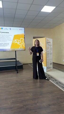 A woman, identified as Oksana Humeniuk, is giving a presentation in a well-lit room with a high ceiling. She is standing to the right of a large screen displaying text and graphics. She is dressed in black, wide-leg pants and a matching short-sleeved top, with a lanyard around her neck. She is gesturing with her right hand as she speaks.

The screen features a title in Ukrainian, "Оксана Гуменюк," and further text below, which appears to be her professional biography and the topic of her discussion panel. The graphics on the screen include yellow abstract shapes and figures. To the right of the woman and the screen is a tall, yellow display board with Ukrainian text, including the words "Миронівка" and "Місто говорить." The floor is a polished wood, and a patterned rug is visible. The setting suggests an indoor event, likely a conference or a public lecture, given the presentation screen and display boards. The overall atmosphere appears professional and informative. The time of day is not explicitly indicated but the lighting suggests it could be daytime. There are no visible weather cues. The location is identified as Myronivka, Ukraine, suggested by the text on the yellow display board.