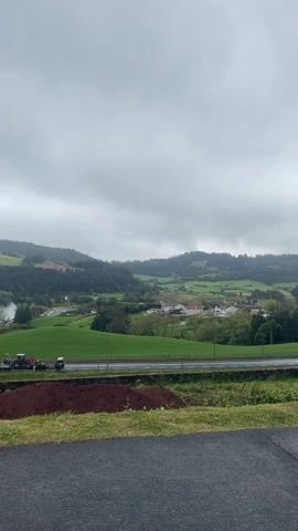 The video shows a landscape view of a rural area on a cloudy day. In the background, there are rolling green hills partially covered in mist. A village with white houses can be seen nestled among the hills. In the midground, there is a road with vehicles, including a tractor, stopped on the side. Closer to the camera is a pile of reddish-brown soil. The foreground consists of a paved road. The sky is overcast. Steam rises from a location in the green fields.