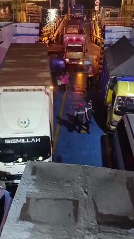 The video shows a nighttime scene on a ferry with several trucks lined up. The perspective is from above, looking down onto the deck. The trucks are parked close together, with some being container trucks and others smaller delivery trucks. There are people on the deck, some wearing reflective vests, seemingly directing traffic or working on the ferry. The deck is wet, indicating it may have been raining or is damp. In the foreground, part of a concrete structure is visible. The background shows lights reflecting on the water and the ramp of the ferry. The overall impression is of a busy loading or unloading operation at night.