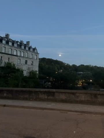 This is a short video clip showing a view of a building and a hill covered in trees. The sky is a deep blue, indicating it is either dusk or dawn, and a bright full moon is visible in the sky. A large, old stone building with multiple windows and a steep, grey roof is on the left side of the frame. The building appears to be old, possibly a historical landmark or apartment complex. In the background, a green, tree-covered hill rises. Lights from buildings and streetlights are visible scattered amongst the trees on the hill, suggesting a town or village. A stone wall or railing runs horizontally across the foreground, partially obscuring the view of the street or pathway below. The road in the foreground is paved and appears to be asphalt, with some cracks visible. The overall atmosphere is tranquil and serene, bathed in the soft light of the moon and the fading daylight.