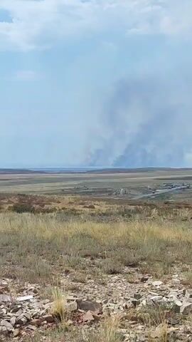 A wide-angle shot captures a vast, open landscape under a partly cloudy sky. In the foreground, dry, sparse vegetation and scattered rocks cover the ground. In the mid-ground, a flatter, grassy plain stretches towards the horizon, dotted with what appear to be some small structures and a road. In the far distance, a large plume of dark gray smoke rises into the sky, indicating a fire. The smoke drifts and expands, obscuring the horizon. The overall scene suggests a rural or wilderness area experiencing a significant fire event.
