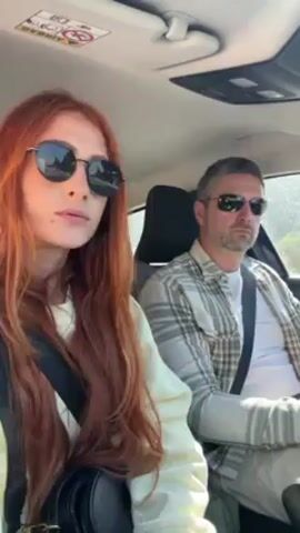 A woman with long red hair and a man are in a car. Both are wearing sunglasses and are buckled in. The woman is in the passenger seat and has a small black purse slung over her shoulder. The man is driving, wearing a plaid shirt. The interior of the car is visible. Music is playing.