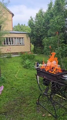 A barbecue grill is set up on a green lawn in front of a light yellow house with white trim. The grill is ablaze with large flames and appears to be cooking food. A garden hose is visible on the lawn. Trees and foliage are in the background. The sky is overcast.