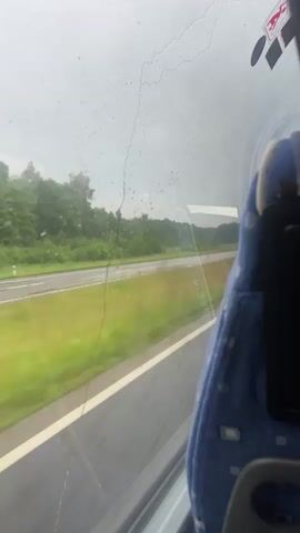This is a first-person perspective video taken from inside a vehicle, likely a bus or train, looking out of a window. The window appears to be wet, with raindrops visible on the glass, suggesting it is raining. Through the window, a landscape is visible, consisting of a road or highway with green grass and trees lining it. The vehicle is in motion, indicated by the blurred motion of the scenery outside. A portion of a blue seat is visible in the foreground, occupying the right side of the frame.