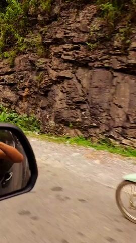 The video shows a car driving on a road surrounded by hills. The car's side mirror reflects a woman taking a photo with a camera. In the distance, there are other vehicles, including a motorcycle. The road is paved and winds through a mountainous landscape covered with lush greenery. The weather appears overcast.