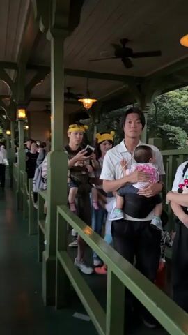 The video shows a queue of people, many of whom appear to be families with young children, waiting in line outdoors. The environment is covered by a roof supported by green posts, with outdoor lighting fixtures visible. People are dressed casually, and some children are wearing character-themed headbands. Several individuals are holding or carrying children. Some people are looking at their phones, while others are interacting with each other or looking ahead in the line. The atmosphere appears to be one of anticipation, possibly at an amusement park or similar attraction. The camera is moving along the line, capturing different individuals and groups as it progresses.