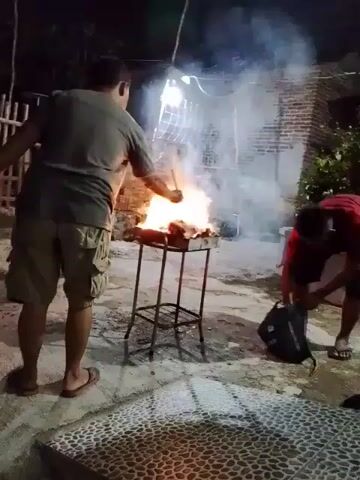 Here is a concise description of the scene:

Two men are outside, apparently grilling meat or something similar on a small, portable barbecue grill. A large flame is visible on the grill, and a significant amount of smoke is being produced. One man is using a flat object to fan the flames, while the other is kneeling near a dark-colored bag, apparently trying to open it. The area is paved with tiles of different patterns. There is a wall in the background. It appears to be nighttime.