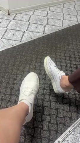 Two pairs of feet wearing white shoes are shown on a floor with mixed surfaces. The floor transitions from a grey and white patterned tile to a dark grey carpet with a square pattern. The left foot is wearing a white sneaker with laces, while the right foot is wearing a white slip-on shoe with small holes. The right ankle is visible, wearing brown pants. The setting appears to be indoors, perhaps a public space due to the mixed flooring types.