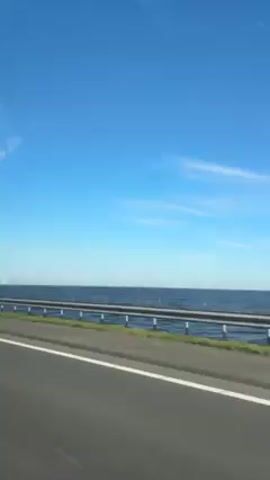 The video shows a road, presumably a highway or interstate, traveling alongside water with a wind farm in the distance. The sky is blue with wispy clouds. A black guardrail separates the road from the water. There's a strip of grass between the road and the guardrail. The wind turbines are numerous and appear as white posts rising from the water. The water is a dark color.