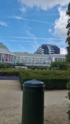 The video shows a view of a botanical garden with a greenhouse and a maze made of hedges. A dark green trash can is in the foreground. The sky is blue with some white clouds and contrails. In the background, there are buildings. The camera is stationary.
