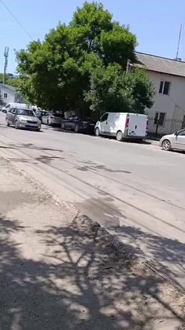 A silver station wagon is driving down a paved street. There are other cars parked on the side of the street. The street is lined with trees and buildings. The sky is clear and blue. The sun is shining.