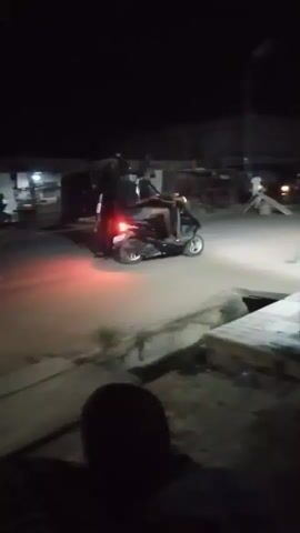At night, a police officer stops a small scooter carrying two passengers on a lit but mostly dark street. The officer, wearing a dark uniform, stands in front of the scooter, holding a flashlight-like object in his left hand and gesturing with his right. The scooter, with its brake lights illuminated, faces the officer. The area includes streetlights, small buildings, and what appears to be a shallow drainage ditch. A person is partially visible in the foreground, seemingly observing the scene. The overall setting seems to be a commercial or residential area at night, with activity occurring under the streetlights.