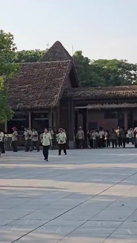 A wide shot captures an outdoor scene in front of a building with a thatched roof. The building appears to be constructed of wood and has several windows and an entrance. Numerous people, many wearing uniforms, are scattered across a paved area. Some are walking, some are standing in groups, and a few are running. The environment appears to be a park or a tourist attraction, with trees visible in the background. The lighting suggests it is daytime, possibly late afternoon given the long shadows.