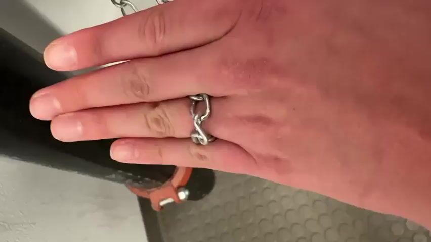 The video shows a close-up of a person's hand wearing a silver chain-link ring on their ring finger. The hand is resting on a black cylindrical object, possibly a pipe, which is secured by an orange clamp. In the background, a white wall and a textured gray floor are visible. The hand then moves away from the pipe, revealing a red object with a chain attached to it, possibly a flushing mechanism. The camera focuses on the ring and the hand as it moves.