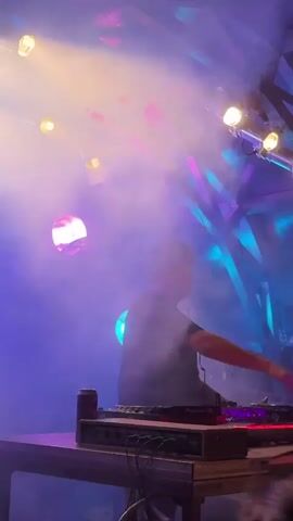 The video captures a DJ performing at a nightclub. The DJ, a male wearing a dark t-shirt and headphones, is seen standing behind a DJ booth. He is operating DJ equipment, including a Pioneer DJ mixer and CDJs. The environment is characterized by dramatic lighting effects, with purple and blue lights illuminating the scene, accompanied by smoke or fog. In the foreground, parts of the audience are visible, with one person's hair and shoulder in focus. A can and other audio equipment are also present on the DJ booth. The DJ appears focused on his performance, occasionally interacting with the equipment. The overall atmosphere suggests a lively event.