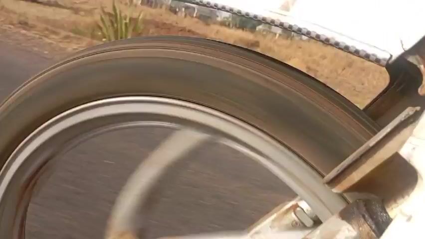 The video presents a close-up view of a wheel in motion, likely on a vehicle. The wheel appears to be made of metal, possibly aluminum, with some visible rust and wear. A brake pad is visible pressing against the wheel, seemingly providing friction. The surrounding structure, likely part of the vehicle's chassis or suspension, is painted white but shows signs of cracking and aging. The background appears to be a blurry landscape, suggesting the vehicle is in motion on a road. There is no visible person in the frame.