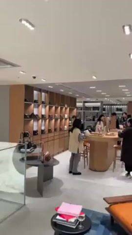 The interior of a luxury retail store is shown. Patrons browse items on shelves and tables. Several store employees dressed in black interact with customers. The interior is well-lit, with wood and soft-colored furnishings. There is a chair with orange upholstery. Clothes hang on racks, and there are shoes on display.