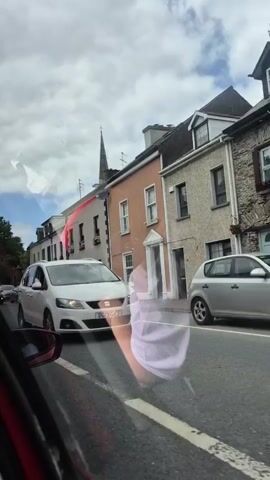 This video captures a street scene in Clonakilty, Ireland, on a cloudy day. The perspective is from inside a vehicle, looking out the window. The foreground shows the side mirror of a red car, with its reflection partially visible. In the middle ground, several cars are stopped or moving slowly in traffic. A white minivan, a white crossover SUV, a dark grey hatchback, and a blue Mini Cooper are among the vehicles. The background features a row of terraced houses with pitched roofs and chimneys, typical of Irish architecture. A tall, narrow spire, likely from a church, dominates the skyline.

A prominent detail is a reflection on the car window, showing a person wearing a light purple garment, possibly a t-shirt or a scarf, with a lighter colored fabric over it. This reflection is quite clear and appears to be of the person filming or someone near them. The lighting suggests it is daytime, but the overcast sky indicates it's not a sunny day. The presence of cars and buildings clearly places the scene in an urban or town setting. The overall mood appears neutral, focusing on the everyday activity of traffic on a street. There is no discernible text visible in the video.