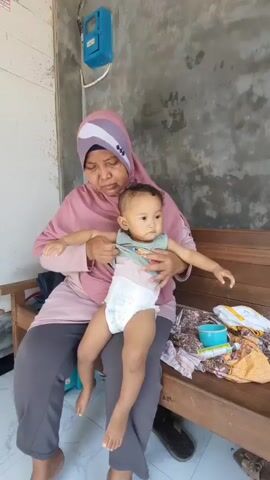 A woman wearing a hijab and pink shirt, sits on a wooden bench, holding a baby on her lap. The baby is wearing a diaper and a light-colored shirt. The woman pulls the baby's shirt over its head, and then pulls a green cloth over the baby's head. The baby is facing the right side of the frame and looking to the right. The background is a wall and a blue electrical box mounted on the wall. To the right of the bench, there are some clothes on the bench.