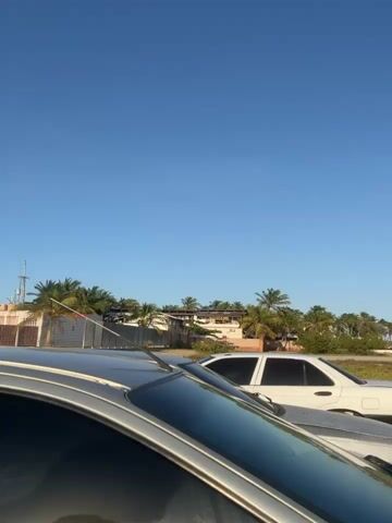 A vertical video shows a parking lot near a beach on a sunny day. At the beginning of the video, there is a silver car and some buildings behind it, including some with palm trees. A woman with long curly hair then walks into the view, walking from the side of the silver car to a black car parked next to it. She walks by the black car. The sun is in the sky, shining brightly. There are other cars in the parking lot. The ground is sandy and looks dry.