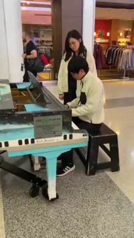 A young man plays a public upright piano with a woman standing behind him. The piano is light blue with black and white keys and is painted with a landscape mural. The man wears a white fleece jacket and dark pants and has dark hair and glasses. The woman also wears a white fleece jacket and dark pants. The woman reaches over his shoulder and places a cell phone on the music stand of the piano. The piano is in a public space, likely a shopping mall, with various storefronts visible in the background and a gray tiled floor. A man with a backpack walks past on the left.