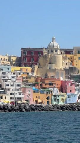 The video shows a coastal view of a town with brightly colored buildings and a church with a dome. The buildings are terraced on a hill that slopes down to a rocky shoreline. The sea is calm and blue, and the sky is clear and bright. The camera is moving slightly, suggesting it was taken from a boat or other moving platform. There are no people visible in the video.