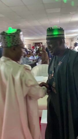 Two men in traditional Nigerian attire are shaking hands indoors. The man on the left is wearing a light pink robe and a green LED-lit cap. The man on the right is wearing a dark green robe with gold detailing and a traditional cap. They are looking at each other and smiling. In the background, people are seated in white chairs and other men are wearing traditional caps. A red carpet is visible on the floor. The room appears to be a large hall or event space. The camera's focus shifts slightly during the handshake.