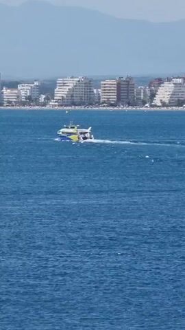 A high-angle shot captures a bright blue ocean. A medium-sized boat with blue and yellow accents is moving from left to right across the water, creating a white wake. In the background, a shoreline is lined with numerous multi-story white buildings. Beyond the buildings, hazy mountains form the distant horizon under a clear, pale sky.