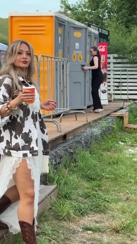 This media file features a group of people at an outdoor event, likely a festival or gathering, in Дрокино, Russia. The foreground is dominated by a woman dressed in a white lace dress with a cow-print jacket and brown cowboy boots, holding a cup and a bag. She appears to be smiling and engaged with the camera.

In the background, other individuals are visible, including two women in swimwear in the middle ground and a man and a woman in the far background, suggesting a social and relaxed atmosphere. The setting includes portable toilets, a white wooden fence, lush green grass, and trees, indicating an outdoor location during daylight hours. The weather appears to be fair, with no visible rain or harsh sunlight.

A prominent sign for Coca-Cola is visible in the middle ground. The overall mood of the scene seems festive and casual, with people enjoying themselves outdoors. The presence of a white canopy structure in the background further suggests an event space. The time of day is likely afternoon, given the lighting.