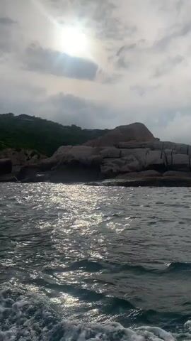 The video shows a view from a boat moving through the water. The water is choppy and reflects sunlight. In the background, there is a rocky coastline covered in green vegetation. The sky is cloudy with some sun visible. The camera appears to be at water level, looking forward and slightly to the side. The movement of the boat is evident by the wake visible in the foreground. There are no people or visible objects other than the natural environment and the boat itself.