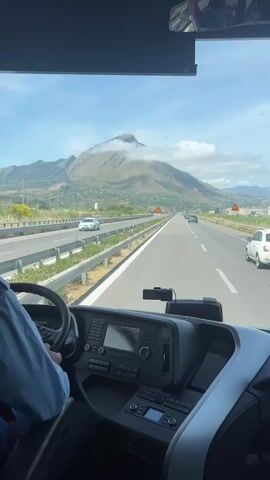 A first-person point-of-view shot shows the inside of a vehicle with a person's hands on the steering wheel and the dashboard in the foreground. The vehicle is traveling on a multi-lane highway with a mountainous landscape visible in the background. The sky is partly cloudy. A speed limit sign indicating 100 km/h is visible on the right side of the road. Other vehicles are also on the highway.